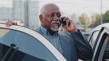 Mature african american businessman standing near car outdoors in parking lot speaks on mobile phone elderly friendly man communicates on telephone answers call using smartphone talking by cellphone