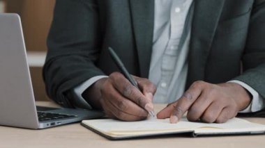 Close-up male hands writing in notebook with pen make notes information financial analysis startup project management idea sitting at desk in office using laptop unrecognizable middle-aged businessman
