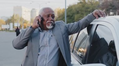 Old african american man stands near automobile communicates on telephone in street parking simple friendly talk carefree elder put hand on car experienced driver gray-haired businessman outdoors