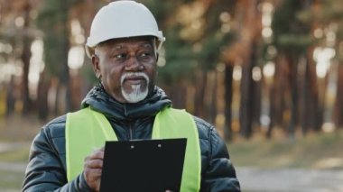 Mature concentrated forestry engineer technician forester in protective helmet stands in park assesses situation makes plan for cutting trees observes reserve writes data takes reforestation measures