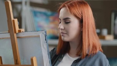 Caucasian woman artist girl painter with red hair enjoying art work with paints creates picture drawing painting on canvas looking at camera smiling posing smile toothy happy cheerful draw laughing