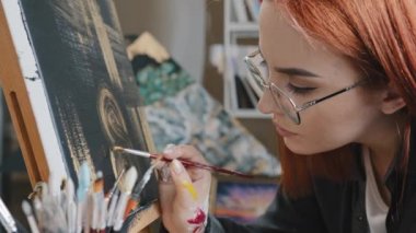 Side view red-haired painter artist creator designer woman student girl in glasses holding brush paints picture on black background with golden oil learning art painting on canvas yellow abstraction