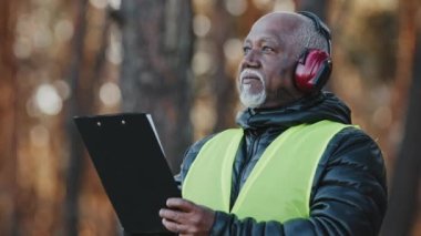 Close-up african american male professional foreman forestry engineer standing outdoors wearing protective headphones assessing situation making tree felling plan writing data reforestation projectt