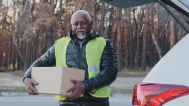 Mature african american male loader worker in uniform outdoors standing near car holding cardboard box elderly gray-haired man in work clothes works in delivery service loads delivers goods parcels