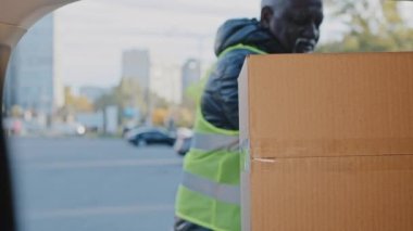 Mature african american man experienced courier loader delivery worker handyman in uniform outdoors putting cardboard boxes with orders in truck car of transport service loading goods in automobile