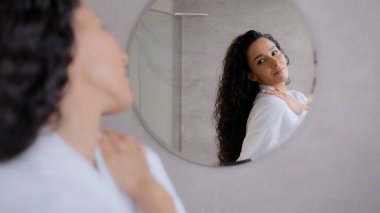Bornozlu genç bir kadın banyoda aynaya bakar. Çekici, kendine güvenen, yansımaya hayran olan, mükemmel fiziğin tadını çıkaran, cilt rengi, doğal güzellik, sabah spa prosedürü.