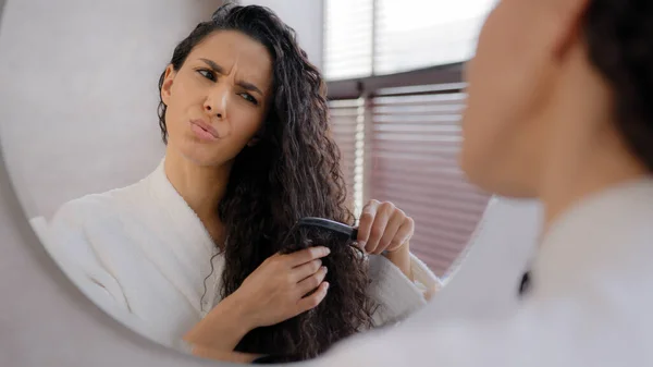 Close-up young woman looking in mirror combing hair in bathroom annoyed sad girl dissatisfied damaged dry tangled hair condition frustrated about split ends vitamin deficiency morning daily routine