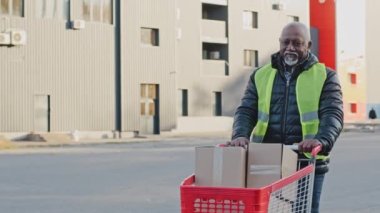 Mature african american deliveryman using handcart with cardboard boxes professional courier loader in uniform transports trolley with parcels to car deliver purchases efficiently delivery service
