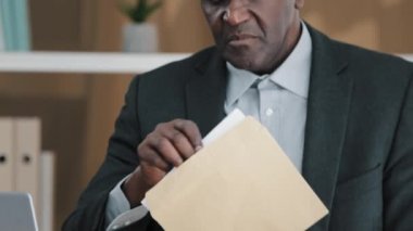 Interested African old 50s middle-aged businessman in office sitting desk with laptop opening envelope reading bad news mail letter feels frustrated sad unhappy high bills dismissal notice bank debt