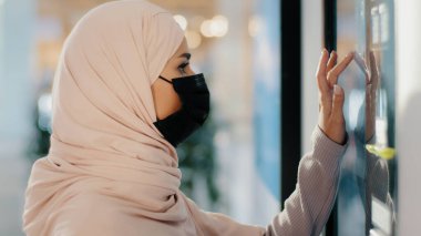 Koruyucu tıbbi maske takan genç Arap kadın self servis makinesinin yanında duruyor. Müslüman kız alışveriş siparişleri alıyor. Bakkaldan servis için ödeme yapılıyor.