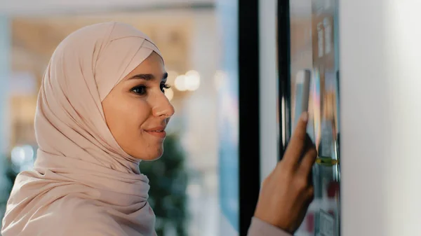 Young happy arab woman in hijab stands near coffee machine orders drink smiling muslim girl making order on self-service automaton pays for service using contactless payment technology on smartphone