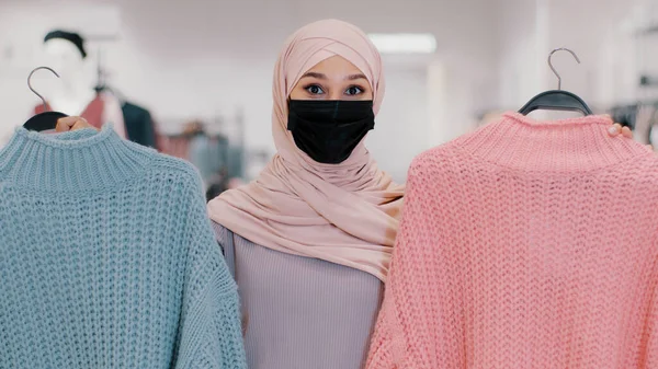 Muslim young woman in medical protective mask during pandemic quarantine islamic girl in hijab seller in clothing store offers sweaters on sale discount female buyer cant choose between two colors