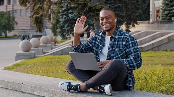 Successful african smiling man guy student male freelancer programmer designer in wireless headphones sitting on terrace in city with laptop working remotely e-learning online waving hello hi hand