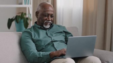 Older bald African American man 60s senior mature businessman use easy wireless technology for comfort work online shopping ecommerce at home couch typing massage chatting banking in computer app