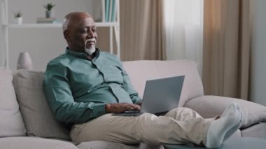African bald overweight obese old middle-aged man senior businessman sitting on sofa at home working with laptop typing feels tired stress relaxing leaning back on sofa pauses after finishing work