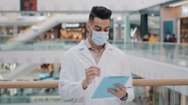 Close up Arabic Indian medical worker man doctor in mask uniform control health safety in public place standing indoors expects writes information appointment expecting writing notes in clipboard