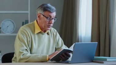 Senior older male online distant teacher webinar speaker tutor coach talking to webcam of laptop video training vlog online conference call old man holding notebook with notes reading information book