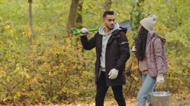 Happy couple young people eco activists volunteers walking in autumn park talking choose suitable place for planting seedling plant tree care about environment social program useful hobby in nature