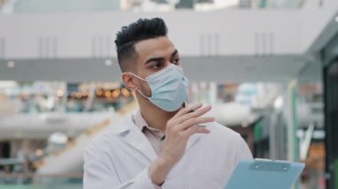 Spanish Arabic Indian worker man doctor in respiratory mask uniform therapist surgeon medical service manager walking in hospital office expects writes information appointment expecting writing notes
