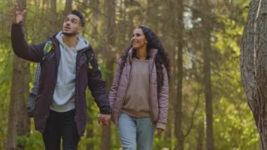 Happy couple in love hikers travel with backpacks walk in forest holding hands hispanic guy and girlfriend enjoy hiking admiring scenery talk about nature tourists walking outdoors enjoying landscape