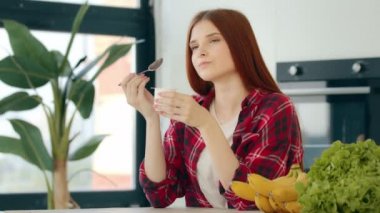 Domestic young woman housewife redhead female eating yogurt with fresh fruits tasting natural milk product dairy dessert use spoon at kitchen enjoying morning breakfast keeping diet healthy nutrition