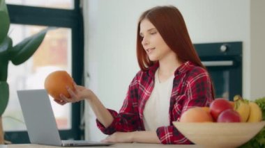 Caucasian young woman food blogger redhead nutritionist sits at kitchen shows at webcam grapefruit citrus recommends online service of delivery products fresh fruits uses laptop video call conference