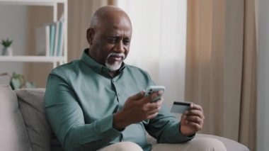 Confused puzzled senior African old bald senior elderly businessman with gray beard has problem with online payment upset retired man error in financial app smartphone money problem transfer at home