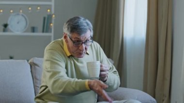 Old elderly mature grandfather man sitting at home couch watching TV drinking hot tea from cup supporting team in online sports competition frustrated dissatisfied with losing failure switches channel