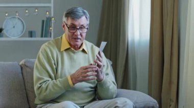 Elderly Caucasian interested man sit couch at home old male receive envelope correspondence grandfather holding paper letter reading good news offer inviting on meeting got bank approval smile happy