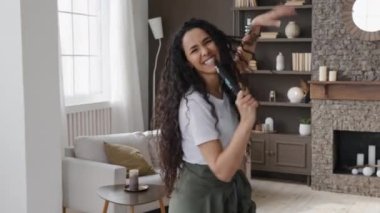 Active funny young woman looking at camera excitedly singing song holding tv remote as microphone having fun at home on weekends amuses dancing vigorously energetic moving to favorite incendiary music