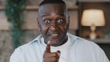 Angry dangerous african american adult senior man looking at camera threatening warning aggressive talking cutting throat with hand show killing gesture of revenge aggression violence standing indoors
