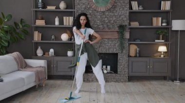 Energetic hispanic woman housewife cleans living room sings song using mop as microphone imagines herself as celebrity on stage attractive girl dancing rhythmic moving to music mopping floor at home