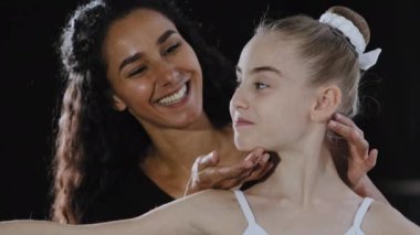 Brunette adult Hispanic woman teacher coach helps teenager girl child rehearsing dance dancing class ballerina learns ballet female trainer corrects posture lifts chin up turns head shows direction