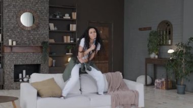 Excited emotion young woman active energetic dancing on sofa singing song using TV remote as microphone happy carefree girl having fun at home alone performing dynamic funny movements moving to music