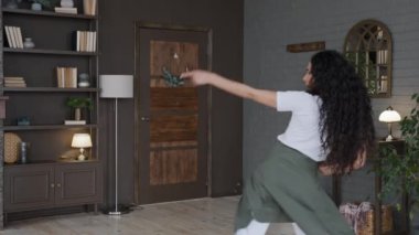 Crazy active young woman dancing in room performs funny move enthusiastic girl with long curly hair having fun at home alone dances to dynamic music energetic merry dance moves rejoices back view