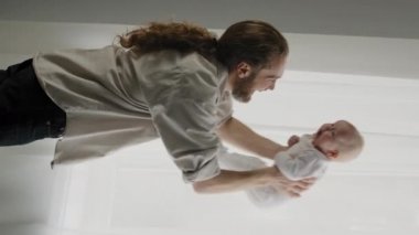 Vertical view Caucasian bearded adult father throw up little cute smiling son daughter infant playing together at home dad holding newborn having fun rising lifting to air throwing kid child catches