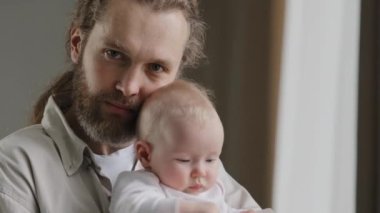Family portrait serious Caucasian bearded dad father man holding hugging small infant newborn girl boy kid daughter son kiss at head looking at camera at home. Closeup daddy kissing adopted child