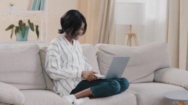 Young woman student freelancer sitting on comfortable couch in cozy room chatting via video call using laptop talking at online conference distant e-learning remote meeting working at home on internet