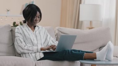 Relaxed female freelancer sitting on comfortable sofa in living room chatting via video call on laptop young businesswoman working remote at home holding business online conference looking at webcam