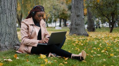 Young focused female student freelancer sitting in autumn park near tree working on laptop typing message checks email studying remote online distance learning using headphones listening to music