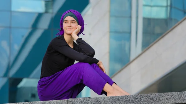 Beautiful calm peaceful girl islamic muslim woman barefoot lady female wears purple hijab sits in city on street building background looking around resting contemplating thinking inspiration dreaming