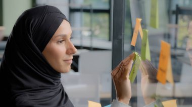 Smiling successful businesswoman corporate leader writing project tasks leaves reminder on sticky colored note organizes work young arab woman office worker in hijab attaches stickers on glass board