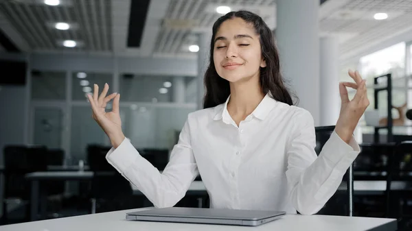 Young tired woman office worker working on laptop typing report finishes work calm balanced female manager meditates with closed eyes resting at workplace improves mental health relaxes practices yoga