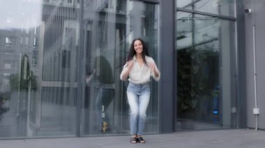 Woman with long curly hair dynamic jumping into air laughing smiling celebrating victory winning rejoicing in success traveling to city standing near wall of building background jump flying having fun