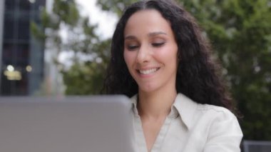 Hispanic Caucasian happy smiling woman university student learning online using laptop computer outdoors, curly hair lady businesswoman smile enjoy remote working job app work with net sitting outside