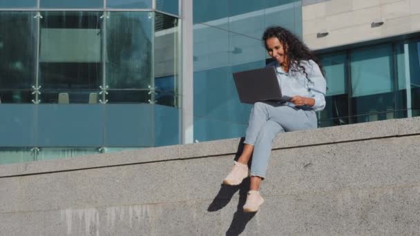 長いショット笑顔幸せな若いビジネス女性フリーランスの女の子学生に座っている通りの街の建物の背景に無線ノートパソコンで勉強オンラインショッピングは良いオファー通知機会を取得します — ストック動画