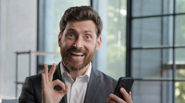 Caucasian bearded businessman 40s boss leader in office with mobile phone victory triumph win victory luck success receive offer money prize opportunity shock ok gesture pointing finger at smartphone