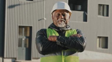 Yakın plan yaşlı Afro-Amerikan inşaat işçisi koruyucu miğfer üniforması içinde açık havada kendine güvenen işçiler göğüs hizasında başarılı müteahhit görünümlü kamera görüntüsü vererek kollarını kavuşturdu.