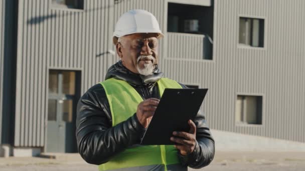老年非洲裔美国人建筑经理评价站在街上戴着防护帽的老年工头 为石板工程师建筑商撰写数据 供他们在室外仔细考虑工作计划 — 图库视频影像