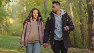 Carefree young hispanic couple in love walks in park holding hands talking guy tells fascinating story happy backpackers travel with backpacks hikers enjoy walking communicate wanderlust concept. High